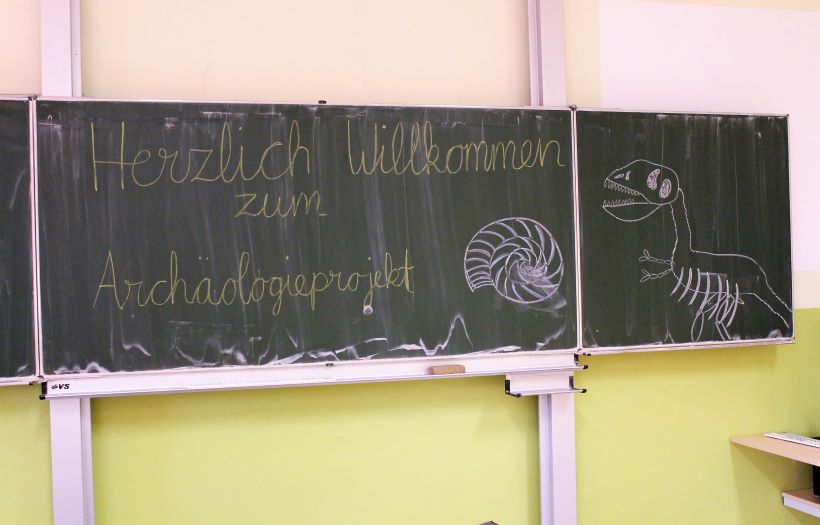 Auf einer Tafel im Klassenzimmer haben Schüler:innen ein Fossil und einen Dinosaurier mit Kreide gemalt. Dazu steht in großen Lettern Herzlich Willkommen zum Archäologieprojekt.