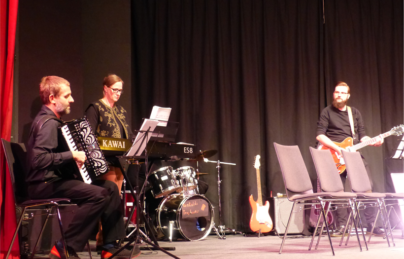 Auf einer Bühne sitzen drei Männer und spielen verschiedene Instrumente.