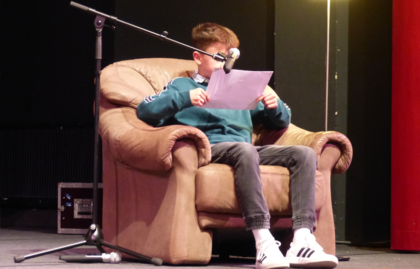 In einem braunen Ledersessel sitzt ein Junge mit einem Buch und liest vor.