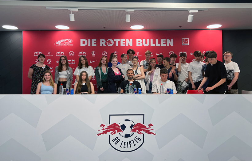 Gemeinsam sitzen Schüler und Schülerinnen mit ihrem Lehrer am Presspult des Leipziger Fußballvereins RB Leipzig.